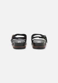 Svarta sandaler med platt sula, som har två breda remmar och en dekorativ spänne. Materialet verkar vara texturerat och syntetiskt.