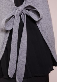 Grå stickad sjal med knytstängning, med mjuk textur och lös passform, över ett svart plisserat lager, som framhäver kontrasterande färger.