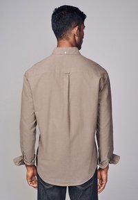 Camicia a maniche lunghe beige chiaro realizzata in tessuto con una finitura testurizzata. Presenta una piega sul retro e un orlo arrotondato. Dettagli del colletto con bottoni.