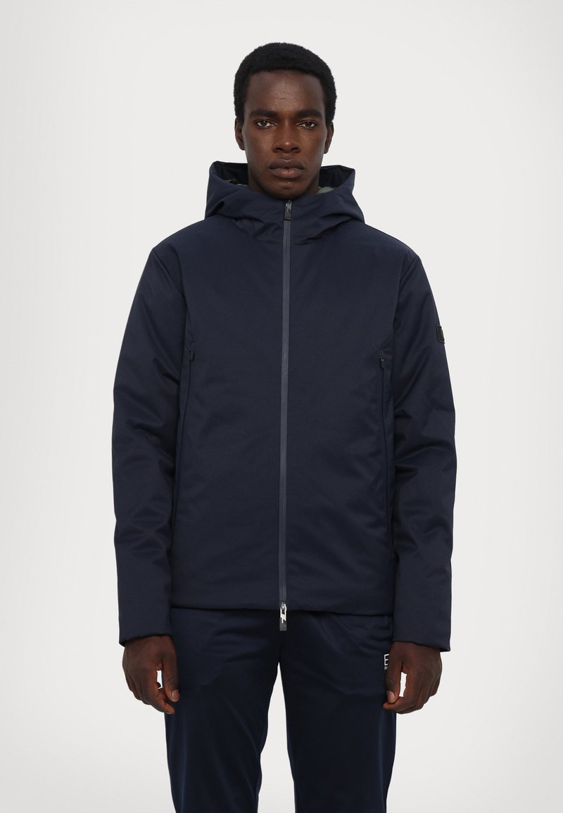Giacca con cappuccio blu navy con chiusura a zip, dotata di tasche laterali e un tessuto liscio e isolante per mantenere caldo. Design semplice, vestibilità moderna.