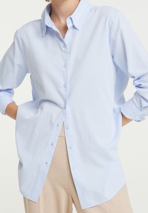 Mujer con camisa abotonada de manga larga color azul claro y pantalones beige, con las manos en los bolsillos, de pie frente a un fondo blanco liso.