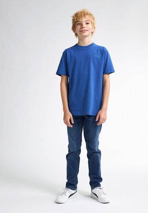 Camiseta de algodón azul con cuello redondo, mangas cortas; combinada con jeans azul oscuro y zapatillas blancas con detalles en negro.