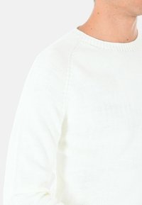 Maglione bianco a maglia con scollo a girocollo, caratterizzato da un motivo testurizzato e maniche raglan. Il tessuto appare leggero e traspirante.
