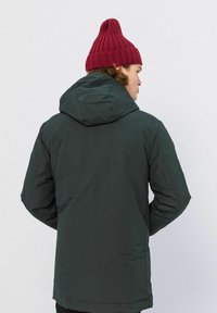 Groene capuchonjack met een gladde textuur en een iets langere achterkant, gecombineerd met een strakke rode ribgebreide muts.