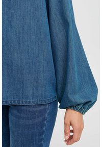 Top in denim blu con vestibilità ampia, maniche lunghe a palloncino raccolte ai polsi e orlo dritto. La texture è liscia con un motivo sbiadito sottile.