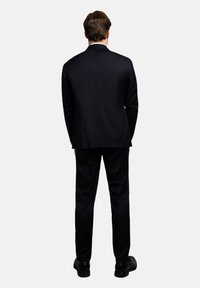 Marineblauer Anzug mit strukturiertem Design, ausgestattet mit einem einzelnen Schlitz und einer geraden Hose, kombiniert mit polierten schwarzen Schuhen.