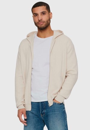 Lys beige glidelås hettegenser med en myk tekstur, frontlommer og snorhette, båret over en hvit t-skjorte og blå jeans.