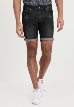 Schwarze Jeansshorts mit umgeschlagenen Säumen und fünf Taschen, ergänzt durch einen roten Akzent an der vorderen Tasche. Kombiniert mit weißen Sneakers.