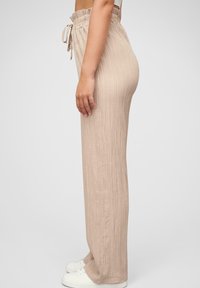 Pantalon large en tissu beige à côtes avec une taille élastique et un détail de cordon, associé à des baskets blanches.