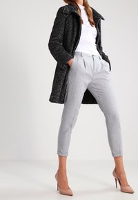 En modell står självsäkert i högklackade skor, iförd en vit topp, grå skräddarsydda byxor och en mörk kappa, poserande mot en ljus bakgrund.