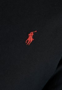 Červené vyšívané logo polo hráče na černé texturované tkanině.