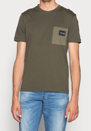 Mężczyzna w oliwkowo-zielonym T-shircie z krótkim rękawem z kontrastową kieszenią na piersi i jasnoniebieskich dżinsach, stoi z rozluźnionymi ramionami.