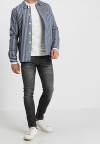Camisa de botones a rayas en azul marino y blanco con puños contrastantes, combinada con jeans grises y zapatillas blancas. Tela suave, corte relajado.