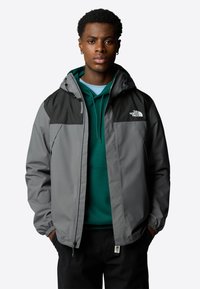 Grijze zip-up jas met een zwart bovenstuk, voorzien van een capuchon en elastische manchetten. Draagt over een groene hoodie, met logodetails op de borst.