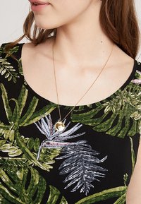 Femme portant un haut noir avec des motifs de feuilles tropicales vertes et violettes et un collier pendentif en or.