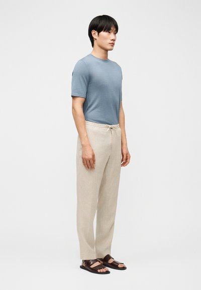 Άνδρας που φοράει ανοιχτό μπλε κοντομάνικο πουκάμισο, μπεζ παντελόνι με κορδόνι και σκούρα καφέ σανδάλια, στέκεται μπροστά σε απλό φωτεινό φόντο.