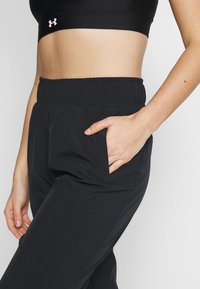 Svarta sportbyxor med elastisk midja, sidofickor och ett mjukt, slätt material. Nära vy av hand i fickan.