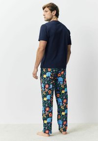 Mann trägt ein navyblaues Kurzarmhemd und dunkle Schlafanzughosen mit buntem Elefanten- und Blumenmuster, läuft barfuß auf einem hellen Teppich.