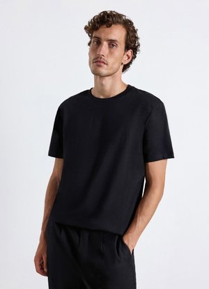 Giovane uomo con capelli ricci che indossa una semplice t-shirt nera a maniche corte e pantaloni neri, in piedi con una mano in tasca su sfondo bianco.