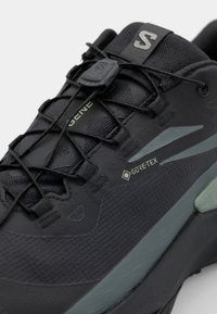 Chaussure de randonnée noire avec tige texturée, logo GORE-TEX et accents verts. Se caractérise par des lacets élastiques et une coque de protection renforcée à l'avant.