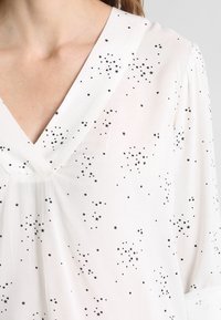 Blusa blanca con cuello en V, confeccionada en tela ligera con un patrón disperso de estrellas negras, mangas cortas y diseño fluido.