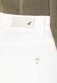 Jeans din denim alb prezintă un buzunar frontat plat cu un accent auriu și un etichetă cu un mic logo de albină la talie. Se vede un top texturat de culoare măslinie.