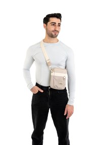 Borsa a tracolla beige realizzata in tessuto resistente, con pattina frontale, tracolla regolabile e hardware semplice. Indossata sopra una maglietta a maniche lunghe azzurra.