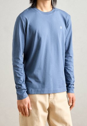Persoon die een blauw shirt met lange mouwen draagt met een klein wit logo op de borst en beige broek, staand tegen een effen achtergrond.