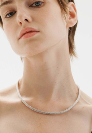 Collar de malla metálica con un diseño liso y redondeado en plata, que se ajusta estrechamente alrededor del cuello, destacando su fina textura y brillo.