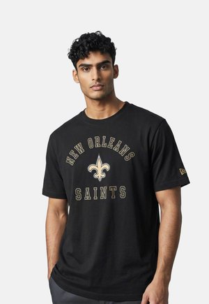Uomo con capelli ricci corti che indossa una maglietta nera dei New Orleans Saints con un logo fleur-de-lis dorato e testo su uno sfondo chiaro uniforme.