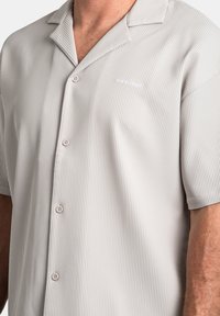 Camicia a maniche corte di colore grigio chiaro con colletto e righe verticali, dotata di bottoni sul davanti e logo del marchio sul lato sinistro. Tessuto morbido.