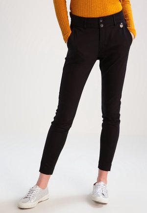 Persona vistiendo pantalones negros ajustados, una camiseta de rayas mostaza de manga larga y zapatillas blancas, de pie con las manos en los bolsillos sobre un fondo blanco.