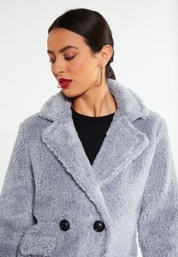 Grå fleecejakke med dobbeltknaplukning, indsnitkrave og sorte knapper. Har et blødt, tekstureret stof for varme.