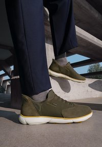 Zapatillas de gamuza verde con suela blanca y acentos amarillos, que presentan un diseño de cordones y detalles de tela texturizada.