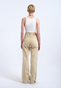 Persoon met kort blond haar, die een witte mouwloze top en beige brede broek met hoge taille draagt, staat met de rug naar voren tegen een effen lichte achtergrond.