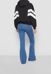 Chaqueta negra con capucha y rayas blancas en las mangas, combinada con jeans de campana azules. El denim tiene un lavado clásico y bolsillos traseros.