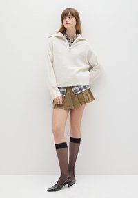 Suéter blanco de punto con cuello tipo mock con cremallera, superpuesto a una camisa a rayas, combinado con una falda verde y medias altas negras. Zapatos de punta con tachuelas.