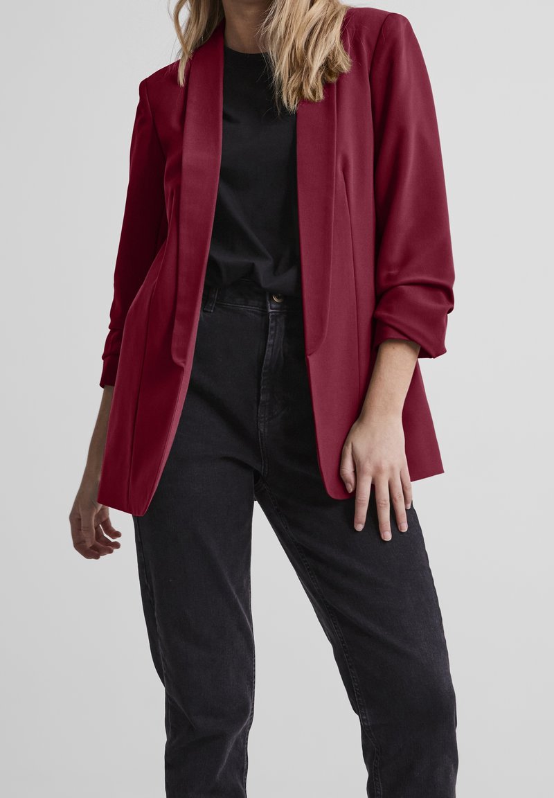 Femme portant un blazer rouge foncé sur une chemise noire et un jean noir taille haute, debout devant un fond gris clair uni.