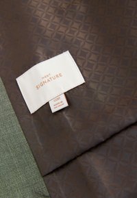 Hvidt tøjmærke med "NEXT SIGNATURE" og størrelse "40R" på brun stof med cirkulært mønster, ved siden af grønt tekstureret stof.