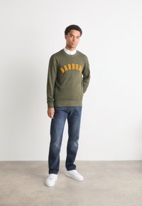 Zelená mikina se žlutými vyšívanými písmeny "BARBOUR", v kombinaci s modrými džíny a bílými teniskami. Hladká textura látky.