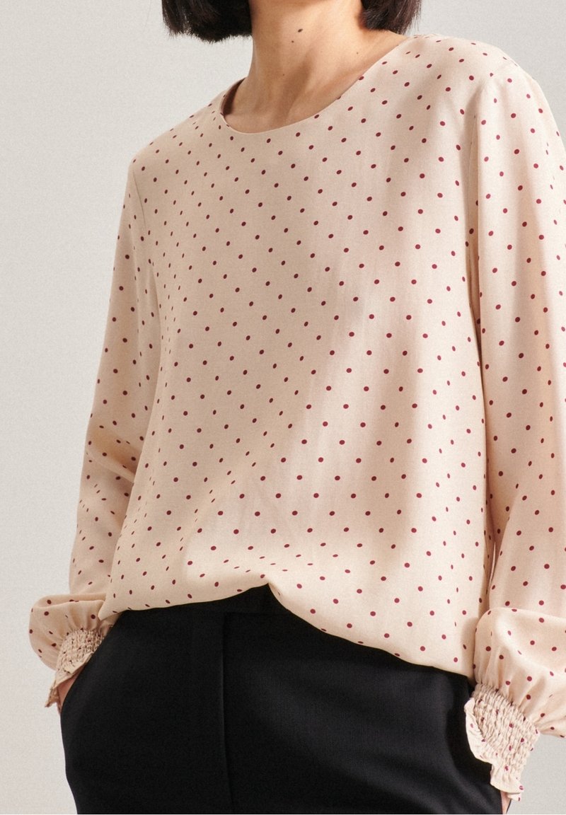 Blusa beige a maniche lunghe con pois rossi, colletto rotondo e polsini elastici. Tessuto morbido con una superficie liscia. Abbinata a pantaloni neri.