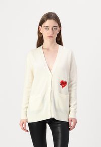 Beige kardián, V nyakkivágással, három gombbal, két elülső zsebbel és piros szív mintával. Puha anyagból készült, bordázott mandzsettákkal és szegéllyel.