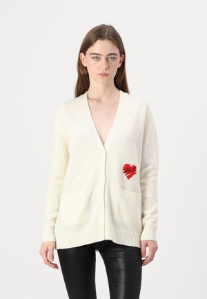 Beige cardigan med V-hals, tre knapper, to frontlommer og et rødt hjerte-design. Laget av myk stoff med ribbede mansjetter og nederkant.