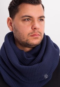 Navy blue knitted scarf, circular design, textural ribbing, features a small fabric logo patch on one side. Soft, warm material.
