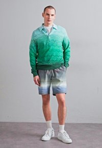 Grüner Gradient-Pullover mit strukturiertem Muster, kombiniert mit kurzärmligen Shorts im Zickzackmuster in Blau und Weiß sowie weißen Sneakers.