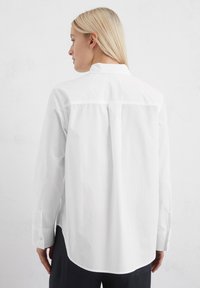 Camicia bianca con bottoni, maniche lunghe, dettaglio della piega sul retro, orlo arrotondato e polsini con bottone; realizzata in tessuto leggero e morbido.
