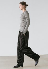 Femme marchant vers la gauche, portant un haut gris à manches longues et un pantalon en satin noir ample, les mains dans les poches, sur un fond uni.