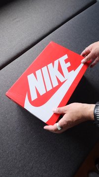 Červená krabička od bot Nike s velkým bílým swooshem a textem, obdélníkový tvar, hladký povrch, držena v ruce na tmavě šedém pozadí.