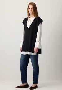 Nő fehér, széles mandzsettás blúzban, fekete ujjatlan V-nyakú pulóverben, kék farmerral és leopárdmintás lapos cipőben, egyszerű fal előtt állva.