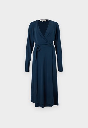 Žena oblečená v tmavomodré dlhšie rukávové zavinovacie šaty so zaviazaným pásom a béžovými sandálmi na vysokom opätku, stojaca na jednoduchom bielom pozadí.